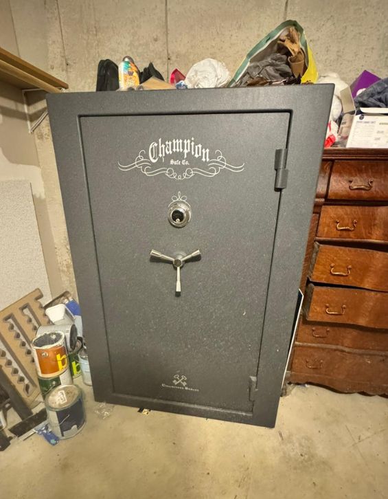 Locksmith in Pacific 63069 1 A large black safe with the "Champion Safe Co." logo is positioned in a cluttered basement, showcasing its sturdy lock and handle. This secure storage solution highlights the importance of professional locksmith services for maintaining safety and access to valuable items.